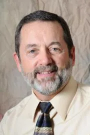 Man with gray beard wearing cream-colored shirt and dark tie smiling at camera
