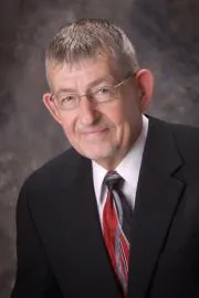 Man with gray hair and glasses wearing black suit jacket and red plaid tie