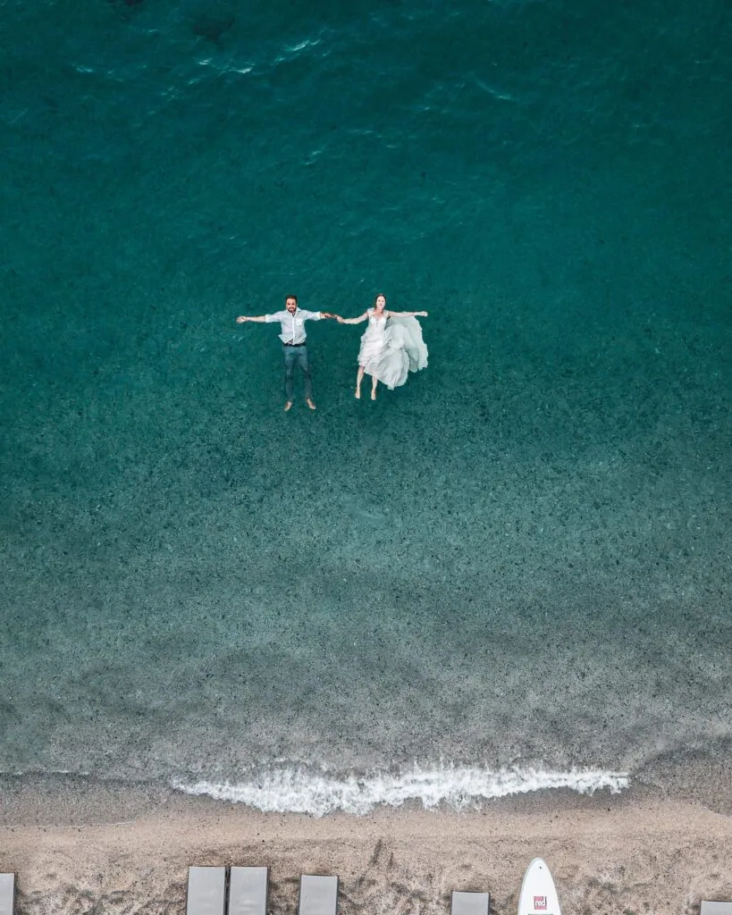Couple holding hands in shallow turquoise ocean water, aerial view from above