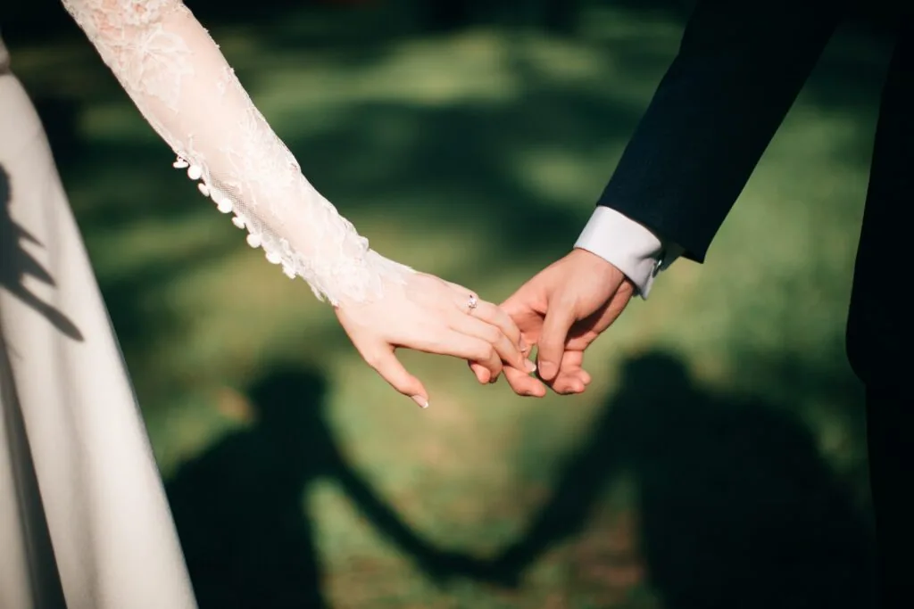 Bride in white lace sleeve and groom in dark suit holding hands outdoors with blurred green background