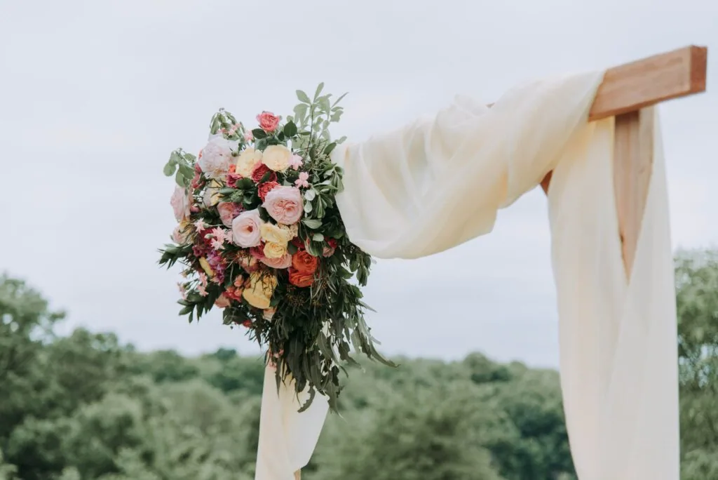 Wedding ceremony arch with cream fabric draping and large floral bouquet featuring pink, red, and yellow roses with greenery, set against outdoor landscape with trees.