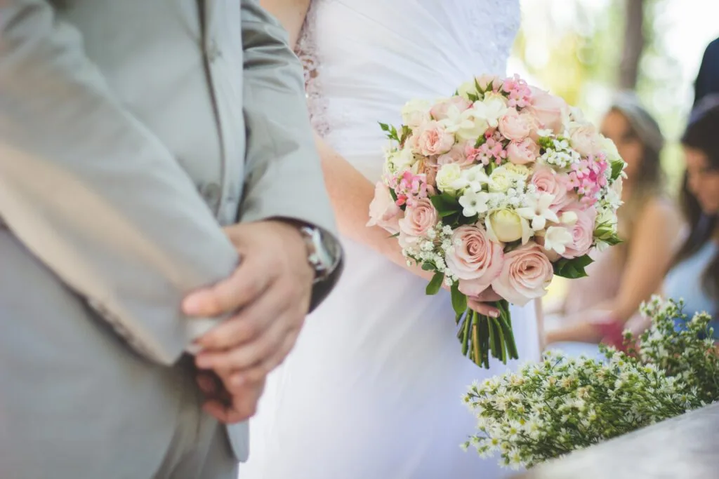 Bride in white dress holding bouquet of pink roses and white flowers standing next to groom in gray suit at outdoor wedding ceremony