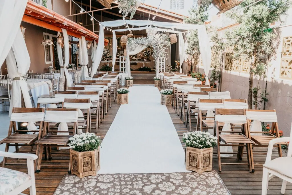 Outdoor wedding ceremony setup with wooden chairs, white aisle runner, white pergola, and potted flowers in a courtyard venue.