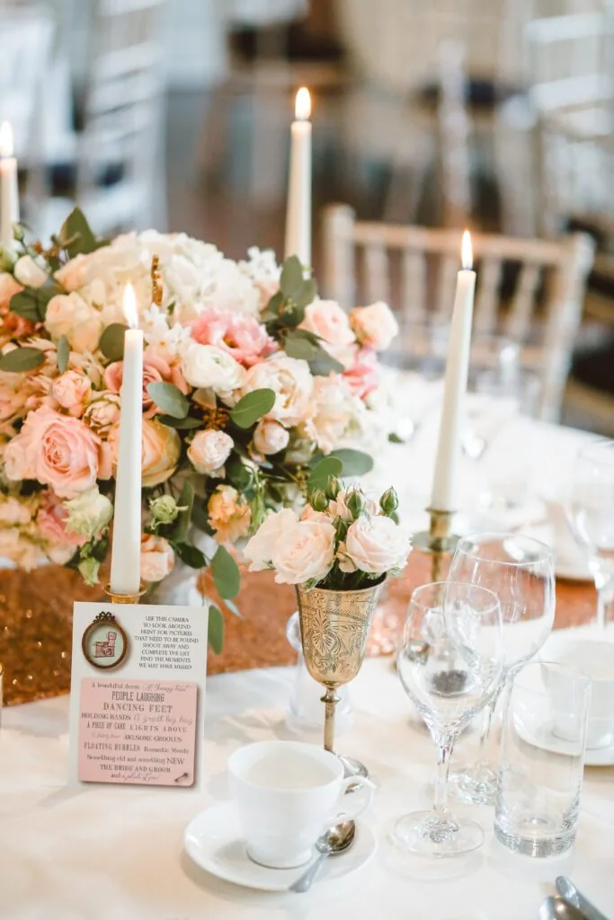 Elegant wedding table setting with blush and cream floral centerpiece in gold vase, white taper candles, place settings, glassware, and pink menu card