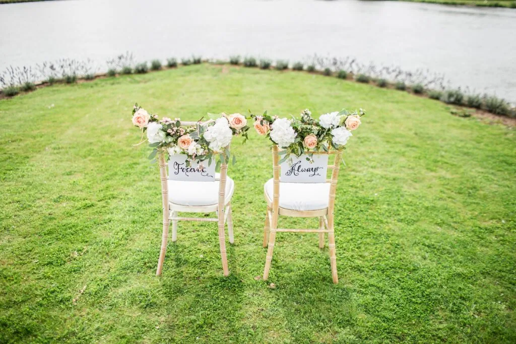 Two wooden chairs on grass decorated with white and pink flowers, facing a water feature with text on the chair backs