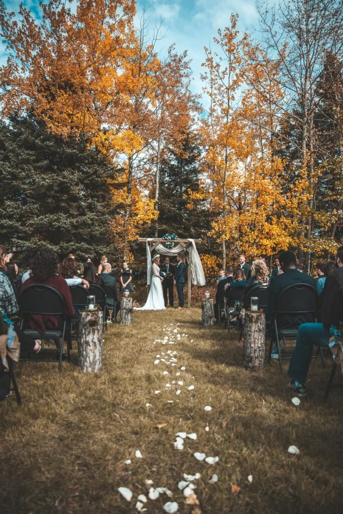 Outdoor autumn wedding ceremony with bride and groom at altar under white arch, surrounded by guests seated on either side, with golden fall foliage trees in background